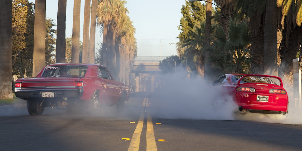 Two way road with red 1967 Chevy Chevelle and bright red MKR Toyota Supra smoking the tires with a huge burnout on either side of the double yellow line in the middle of the road