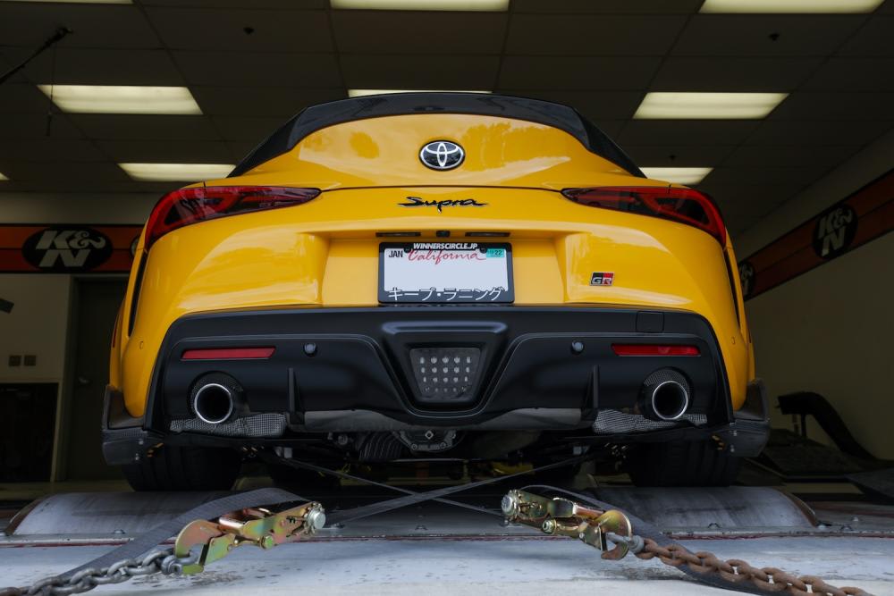 Low angle view of a bright yellow 2022 Toyota Supra GR with wide rear tires, a carbon fiber diffuser and spoiler as well as large exhaust tips on either side secured to the K&N dyno to measure how much horsepower it's making